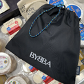 Black BYBBA drawstring bag with blue-yellow rope handle, packable, foldable, reusable, made from recycled ocean plastic.